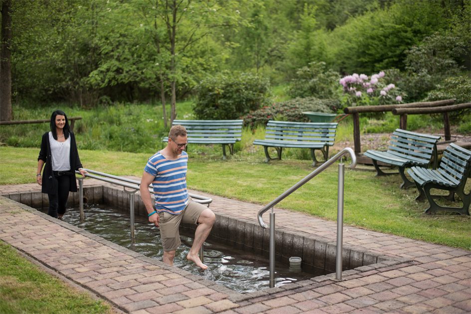 Ein Kneipp-Fußbad mit Handläufen, umgeben von grünen Bäumen und Parkbänken.
