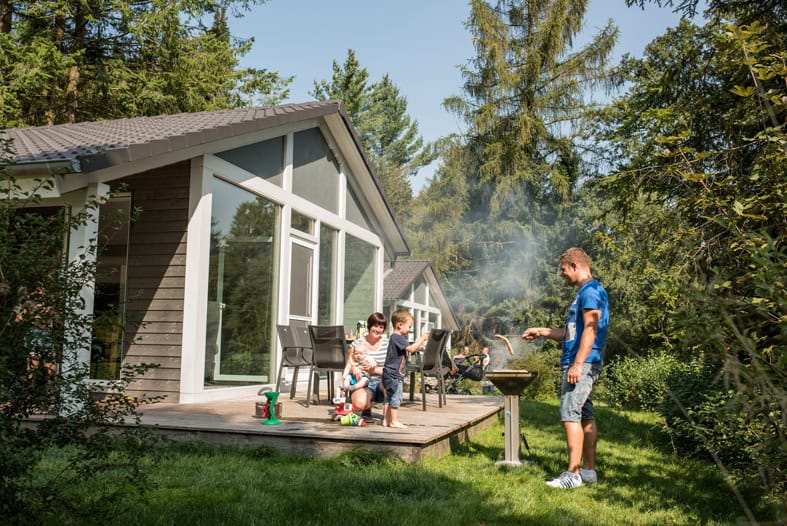 Eine Familie grillt vor einem Ferienhaus im Grünen und genießt die Ruhe.