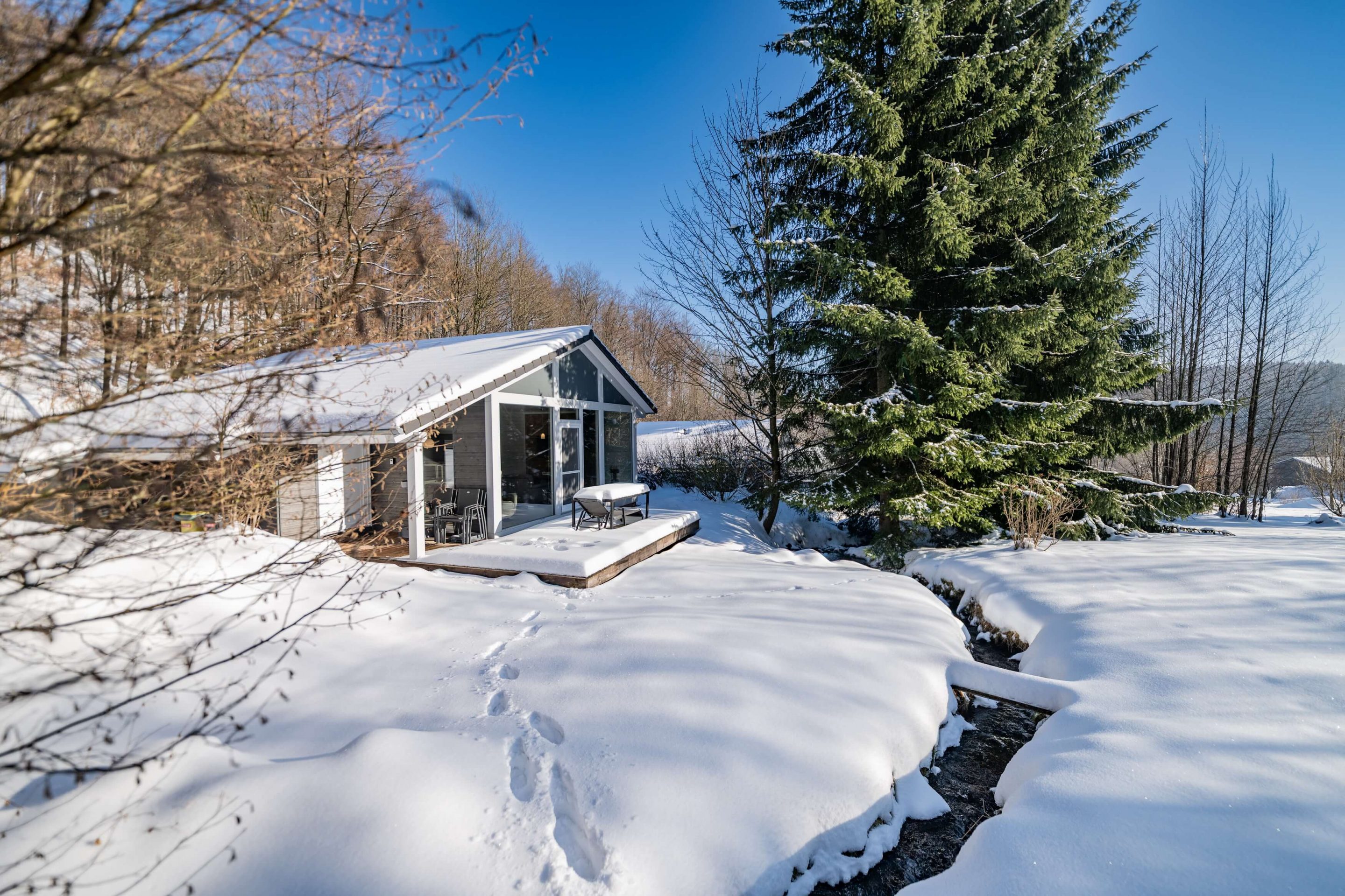 Die Aufnahme zeigt das Ferienhaus von der Seite, wobei die verschneite Terrasse mit Gartenmöbeln und die großen Fensterfronten im Fokus stehen. Ein kleiner Bach schlängelt sich durch die tief verschneite Gartenanlage im Vordergrund, in der deutliche Fußspuren im Schnee zu sehen sind. Eine mächtige, schneebedeckte Tanne flankiert das Haus, während der blaue Himmel für eine helle, freundliche Winterstimmung sorgt.