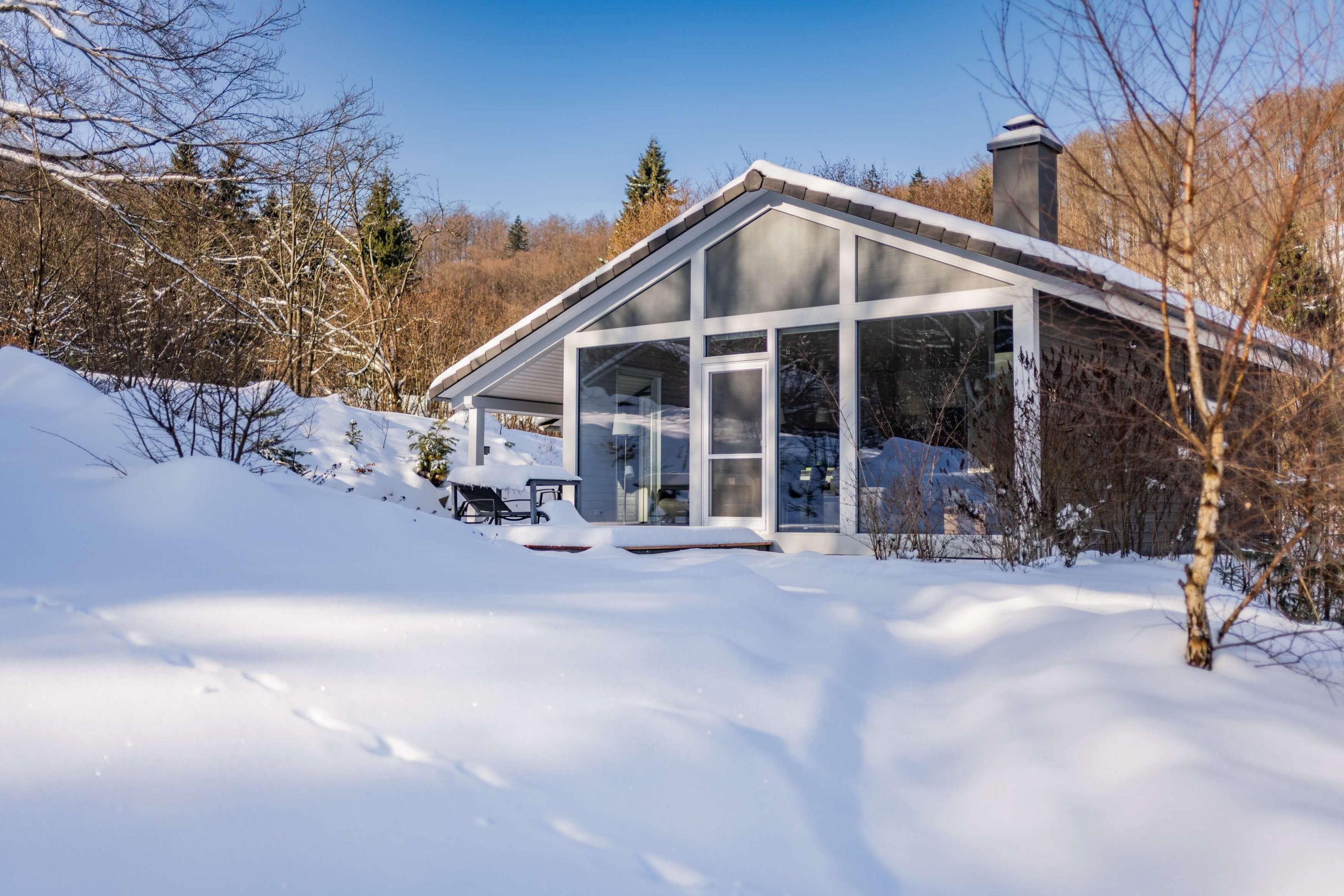 Ein modernes Ferienhaus mit markanter Giebelfront und großzügiger Verglasung steht inmitten einer unberührten, dicken Schneedecke. Im Hintergrund erstreckt sich ein bewaldeter Hang, während die tiefstehende Wintersonne lange Schatten auf den Schnee wirft. Rechts am Haus ist ein dunkler Schornstein sichtbar, und im Vordergrund zeigen sich vereinzelte kahle Zweige im Sonnenlicht.