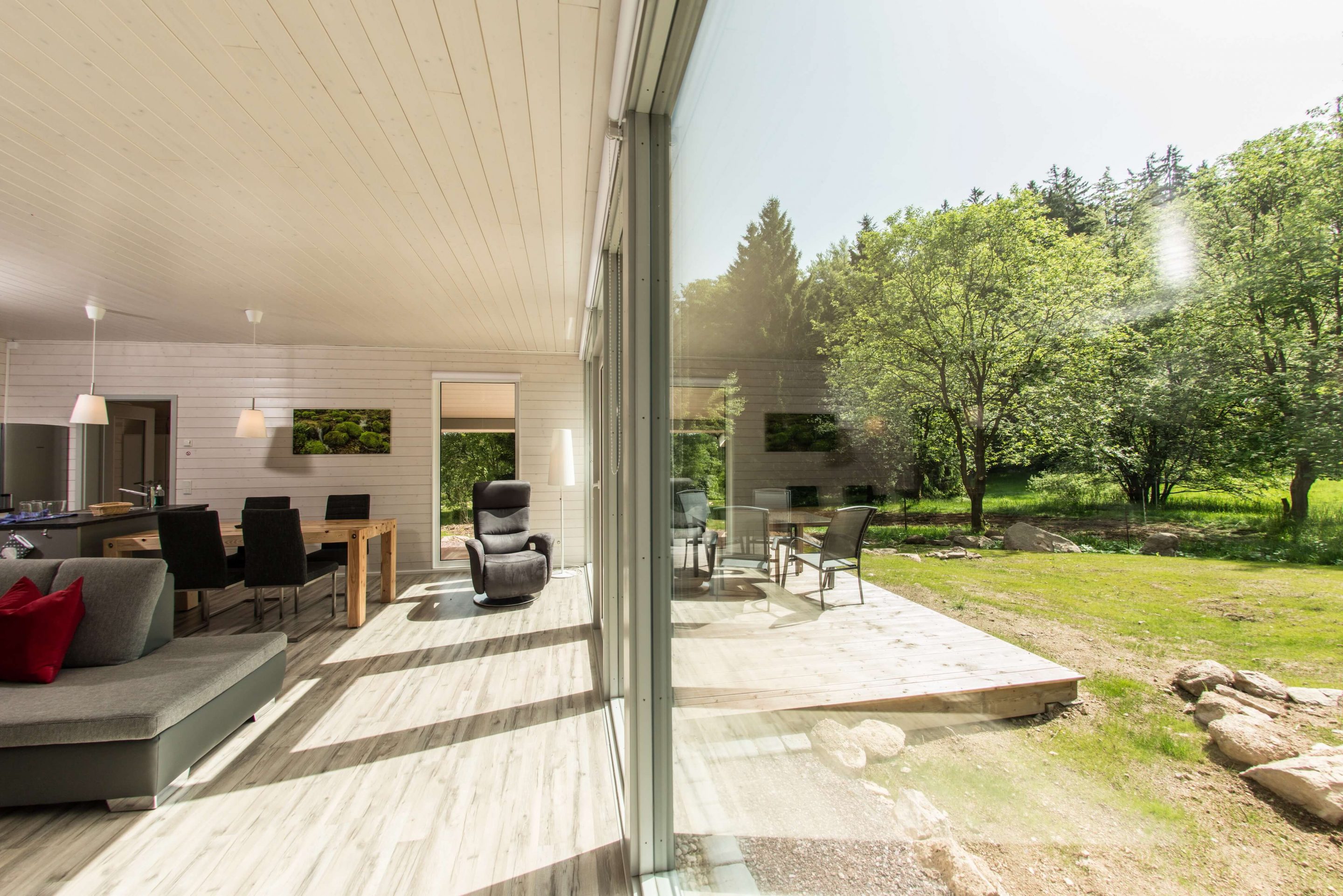 Helles, modernes Wohnzimmer mit hellen Holzwänden, einem grauen Sofa, einem Esstisch, einem Liegesessel und einer Glaswand, die auf eine Terrasse mit Bäumen führt