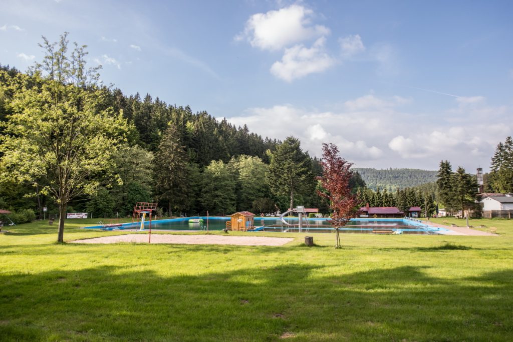 Freibad im Grünen mit Schwimmbecken, Rutsche und Sprungturm vor einer Waldkulisse