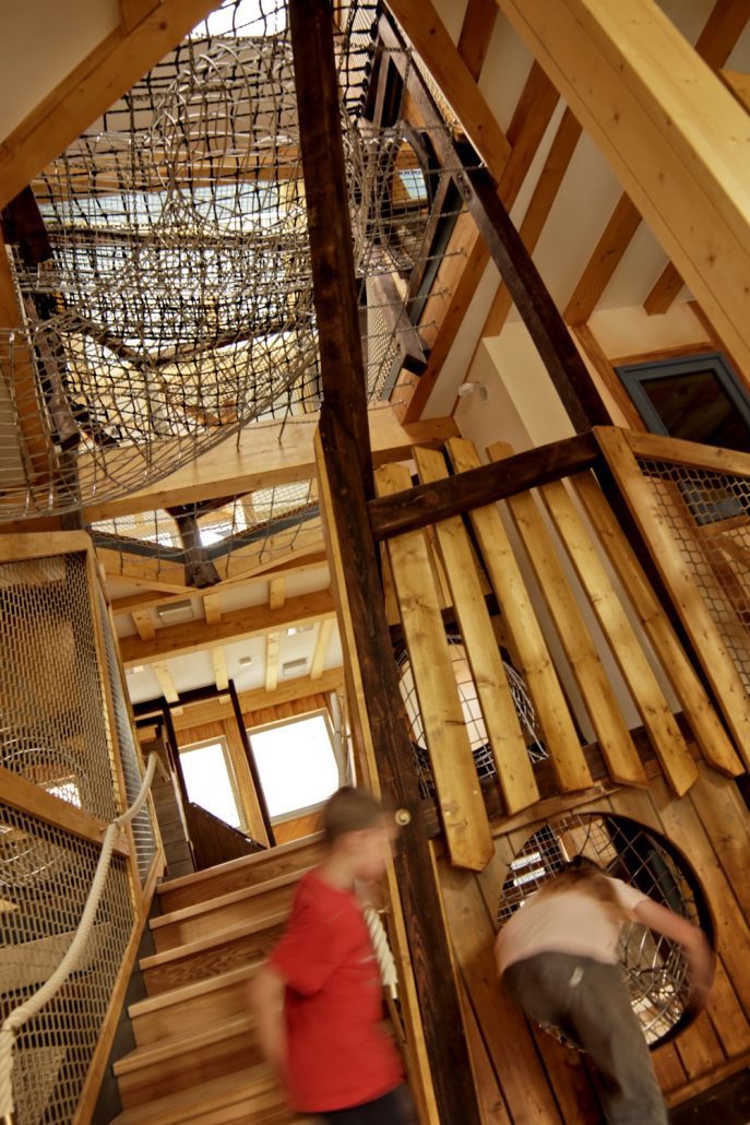 Innenansicht einer mehrstöckigen Spielanlage aus Holz mit Netzen, Treppen und zwei spielenden Kindern, die unscharf abgebildet sind