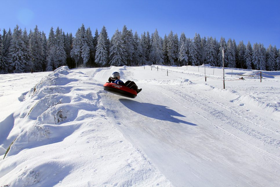 Kind beim Snowtubing auf einem verschneiten Hügel mit Kiefern unter strahlend blauem Himmel